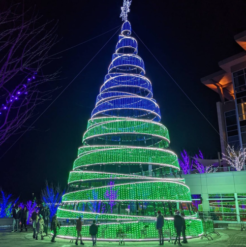 A brightly lit Christmas tree with green and blue lights, surrounded by people enjoying the festive atmosphere at night.